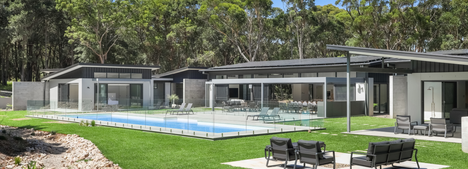 Modern luxury home featuring COLORBOND® steel in Monument color, with LYSAGHT® KLIP-LOK 700 HI-STRENGTH® roofing and ENSEAM® cladding. The property includes a sleek glass-fenced swimming pool, lush green lawn, and spacious outdoor entertaining areas with contemporary lounge seating.