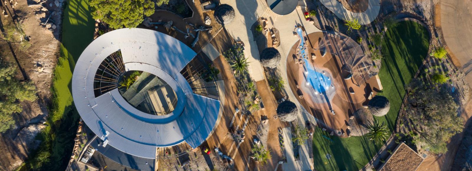 Aerial view of a modern architecture project featuring a curved steel roof, landscaped pathways, greenery, and a nature-inspired playground. 