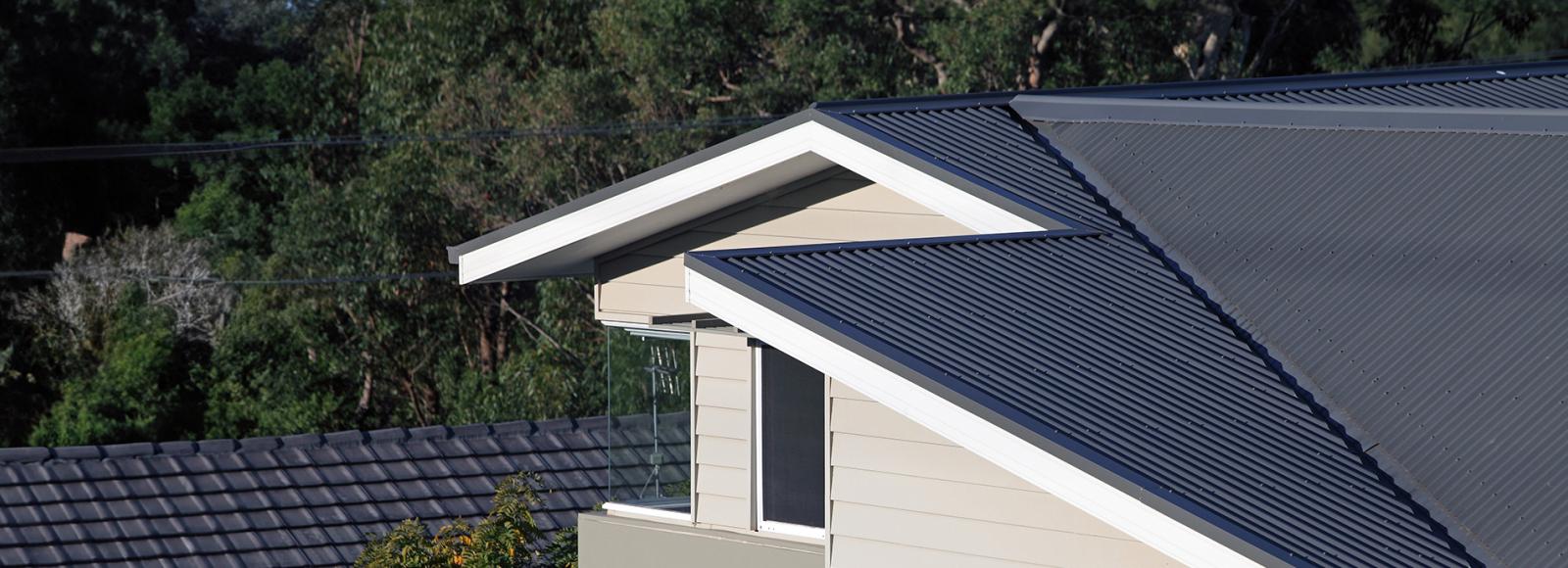 Modern house roof with dark grey corrugated metal sheets and white-trimmed gable design. The exterior walls are light beige with horizontal siding. 