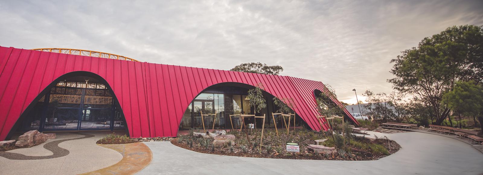 A modern building featuring a bold red facade made of KLIP-LOK 700 HI-STRENGTH® metal sheeting, with distinctive curved archways. Large glass windows and doors reflect the surrounding trees and landscape. 