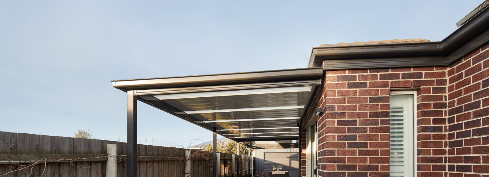 Modern brick home with covered outdoor patio, gable roof carport with polycarbonate panels, and concrete driveway in landscaped backyard