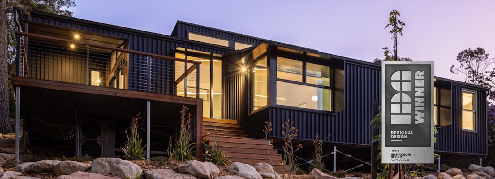 Blackheath Mountain Retreat at dusk, black steel cladding and lit timber decks, with a Regional Design Winner badge.