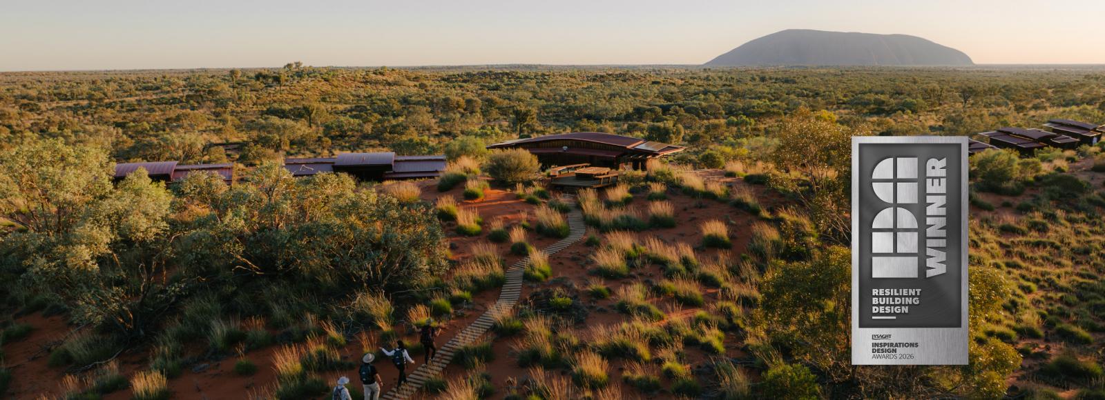 Aerial view of the eco-lodges among red dunes and spinifex, Uluru on the horizon, with a Resilient Building Design badge.