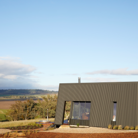 Farmstay featuring a striking angular design, constructed with COLORBOND® steel and CUSTOM ORB® steel solution by Lysaght. The exterior showcases dark, vertically clad steel panels complemented by a sleek, durable metal roof with solar panels. Set against a picturesque rural landscape.