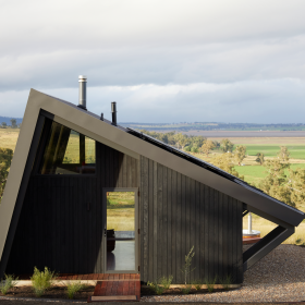 Farmstay featuring a striking angular design, constructed with COLORBOND® steel and CUSTOM ORB® steel solution by Lysaght. The exterior showcases dark, vertically clad steel panels complemented by a sleek, durable metal roof with solar panels. Set against a picturesque rural landscape.