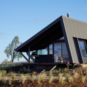 Farmstay featuring a striking angular design, constructed with COLORBOND® steel and CUSTOM ORB® steel solution by Lysaght. The exterior showcases dark, vertically clad steel panels complemented by a sleek, durable metal roof with solar panels. Set against a picturesque rural landscape.