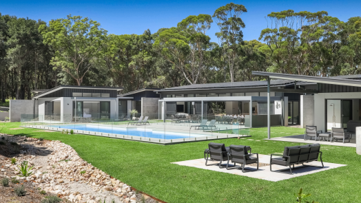 Modern luxury home featuring COLORBOND® steel in Monument color, with LYSAGHT® KLIP-LOK 700 HI-STRENGTH® roofing and ENSEAM® cladding. The property includes a sleek glass-fenced swimming pool, lush green lawn, and spacious outdoor entertaining areas with contemporary lounge seating.