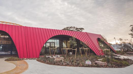 A modern building featuring a bold red facade made of KLIP-LOK 700 HI-STRENGTH® metal sheeting, with distinctive curved archways. Large glass windows and doors reflect the surrounding trees and landscape. 