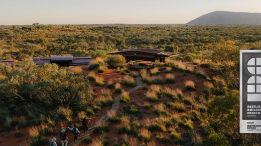 Aerial view of the eco-lodges among red dunes and spinifex, Uluru on the horizon, with a Resilient Building Design badge.
