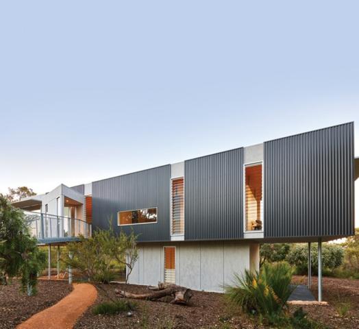 Modern house with a dark grey angular facade, elevated on stilts, featuring orange window frames and a curved orange pathway leading to it, set against a light blue sky.