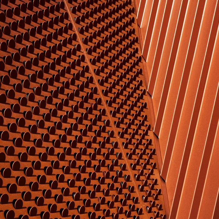 Close-up of ochre steel screening with circular openings beside ribbed wall cladding.