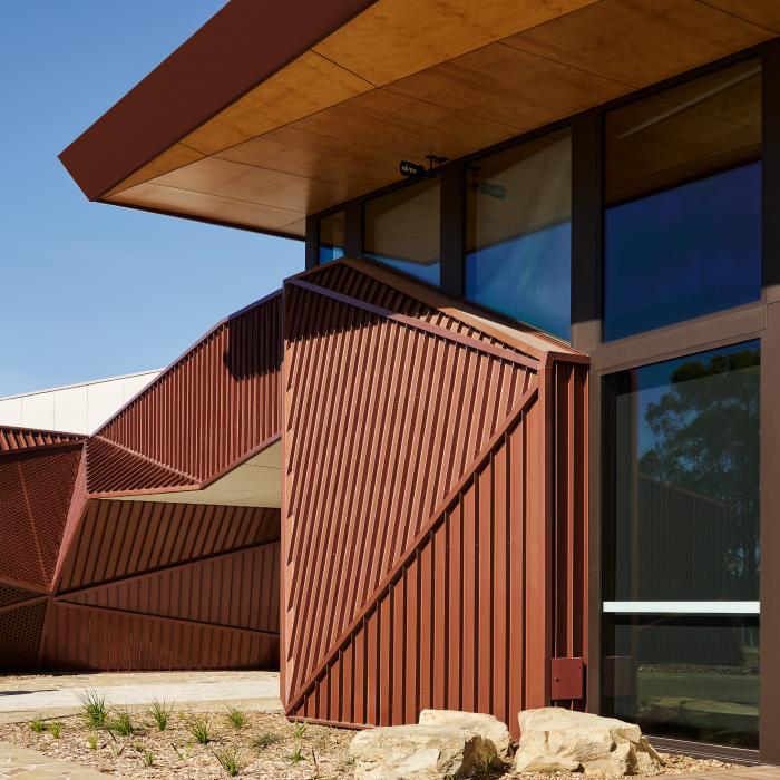 Folded ochre steel walls beside tall windows beneath Larnangurrak’s timber-lined roof.