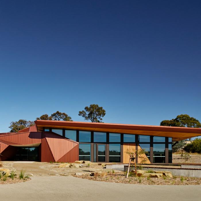 Larnangurrak’s glazed facade and folded ochre walls beneath a broad roof, with native planting.