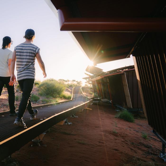 Two people walk along an elevated walkway beside a steel-clad lodge at sunset