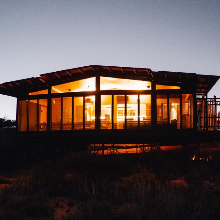 An eco-lodge glowing with warm interior light at dusk, raised above desert scrub
