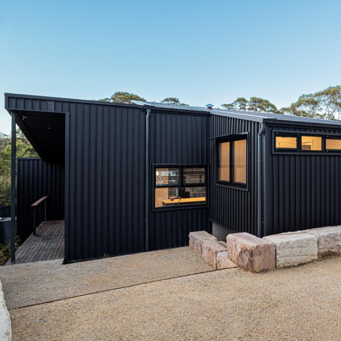 Low mono-pitched forms clad in black steel, with warm light showing through small windows.