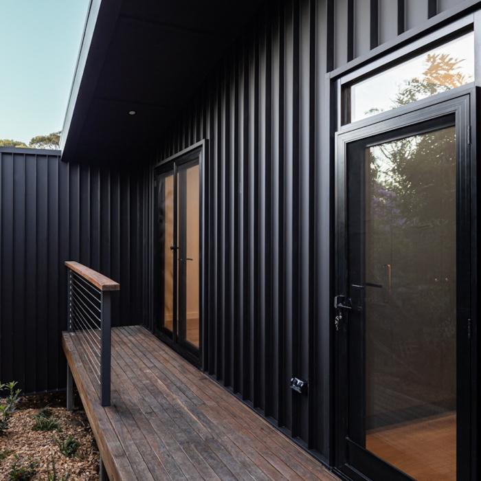 A narrow timber deck runs beside black ribbed cladding and glazed doors reflecting the trees.