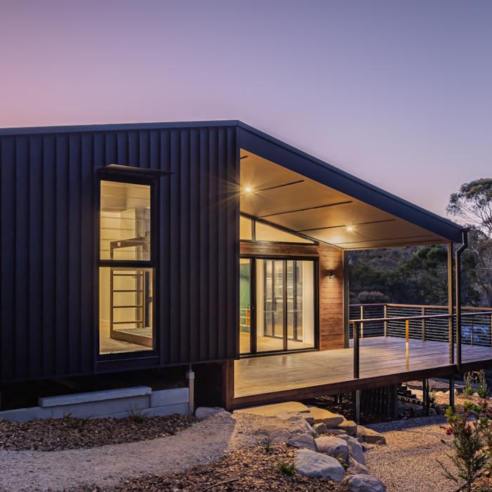 Corner of the retreat at dusk, its raked roof sheltering a lit deck above a gravel and boulder garden.