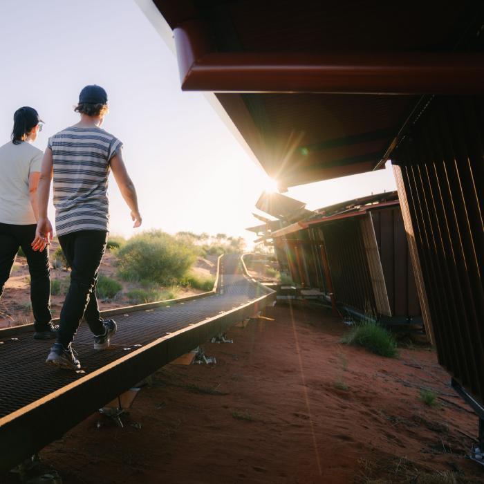 Two walkers on a raised boardwalk beside a lodge with a deep red roof overhang, the sun low behind them.