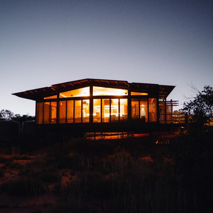The main lodge glowing at dusk, full-height glazing beneath a broad, floating roof above the dark desert.