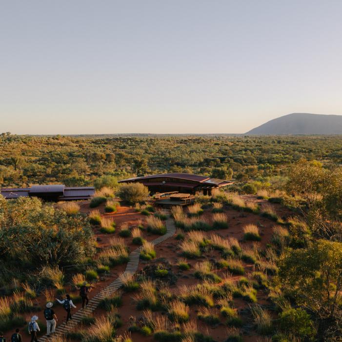 Walkers approach desert lodges at Uluru-Kata Tjuta Signature Walk, with Uluru on the horizon.