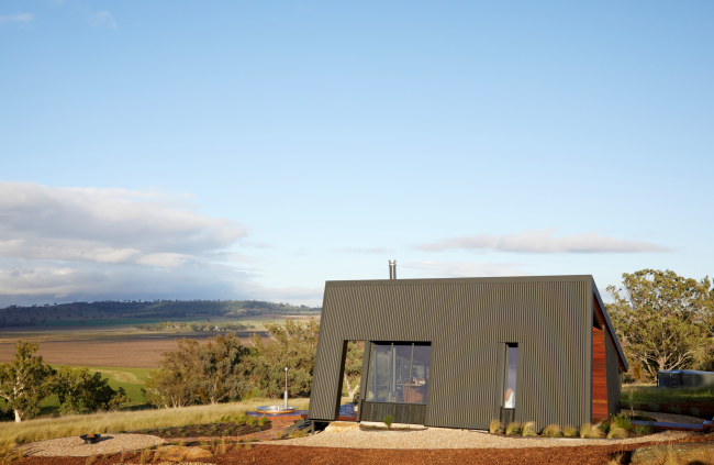 Farmstay featuring a striking angular design, constructed with COLORBOND® steel and CUSTOM ORB® steel solution by Lysaght. The exterior showcases dark, vertically clad steel panels complemented by a sleek, durable metal roof with solar panels. Set against a picturesque rural landscape.