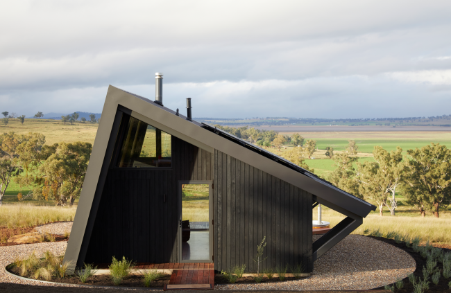 Farmstay featuring a striking angular design, constructed with COLORBOND® steel and CUSTOM ORB® steel solution by Lysaght. The exterior showcases dark, vertically clad steel panels complemented by a sleek, durable metal roof with solar panels. Set against a picturesque rural landscape.