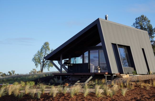 Farmstay featuring a striking angular design, constructed with COLORBOND® steel and CUSTOM ORB® steel solution by Lysaght. The exterior showcases dark, vertically clad steel panels complemented by a sleek, durable metal roof with solar panels. Set against a picturesque rural landscape.