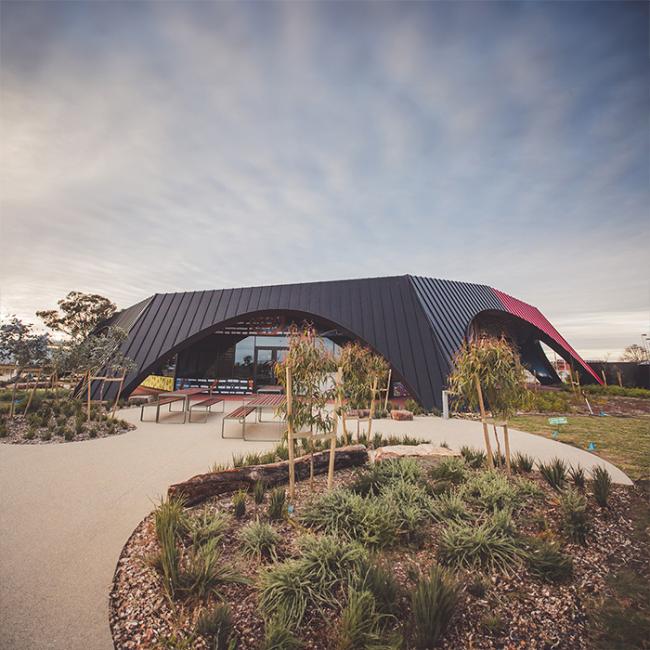 A modern building with a sleek, curved black roof made from KLIP-LOK 700 HI-STRENGTH®, nestled among landscaped gardens and pathways under a cloudy sky.